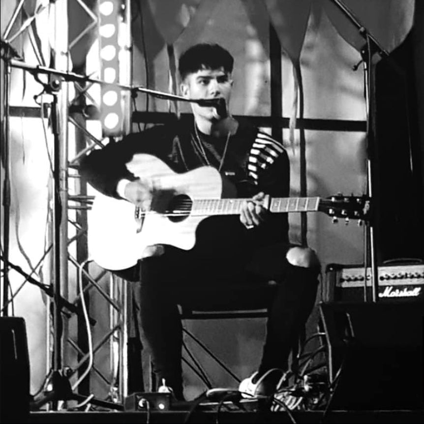 A black and white photo of a male musician sitting on a stage, holding an acoustic guitar and singing into a microphone. Stage equipment, lighting, and amplifiers are visible in the background.